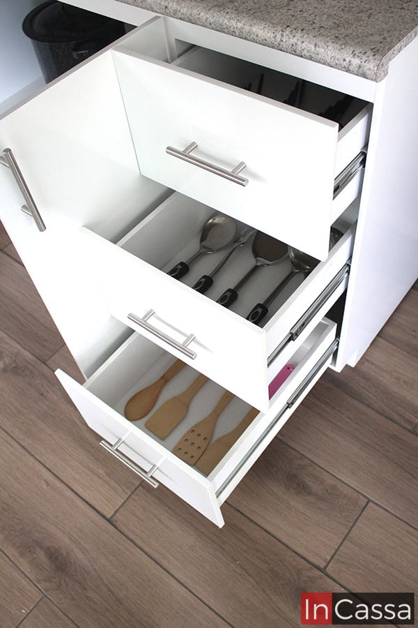 Imagen de los cajones incorporados al módulo de tarja de la cocina, los cuales se muestran abiertos dejando ver la estructura completa de cada compartimento así como el espacio que ofrecen para guardar utensilios. Cada cajón es completamente blanco (incluso en sus interiores), y cuenta con un sistema de rieles metálicos para su apertura y cierre, complementado por una jaladera de barra T instalada al doble frente de cada compartimento.