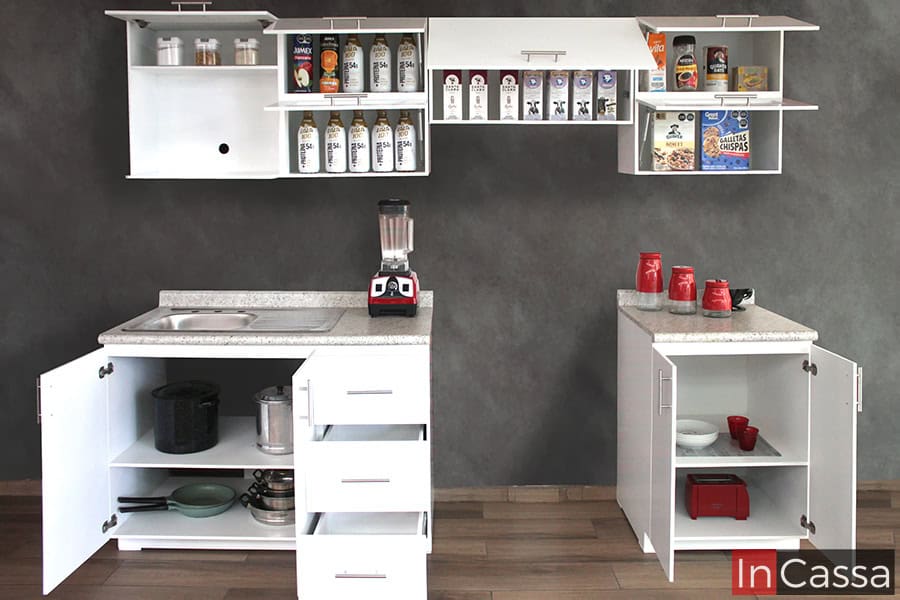 La foto presenta los cinco módulos que conforman la cocina, estos son mostrados con sus puertas y cajones abiertos dejando a la vista todos sus compartimentos. Como puede notarse en la toma, todos los módulos mantienen un vistoso color blanco en la totalidad de su estructura.