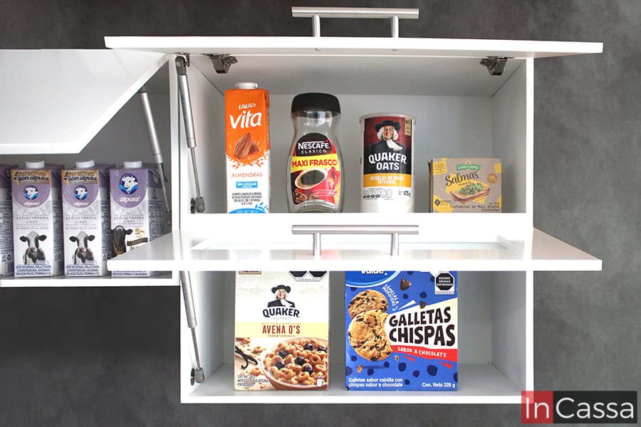 Una foto del módulo sobre carnicero de la cocina, en la que también se tiene una vista parcial del mueble sobre campana. Ambos módulos se muestran con sus puertas abiertas, dejando visibles los pistones neumáticos que dan una apertura y cierre vertical a cada puerta; cada compartimento es decorado con algunos consumibles con los que se puede determinar la capacidad de cada espacio. El interior de cada compartimento mantiene el mismo tono blanco que la estructura externa de los módulos.