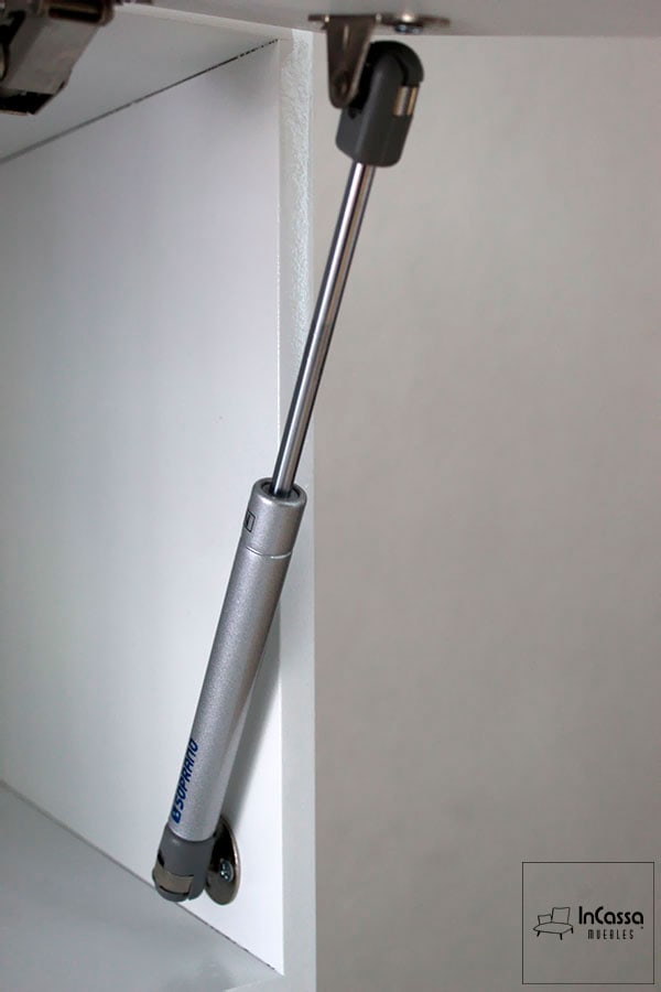 La imagen muestra en primer plano un pistón neumático de gas instalado en uno de los módulos superiores de la cocina AUSTRIA. Este herraje de alta calidad garantiza una apertura suave y controlada de las puertas abatibles, complementando perfectamente el acabado blanco del mueble.