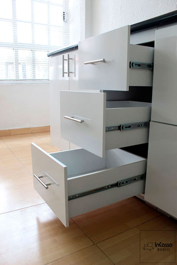 Una toma lateral muestra los tres cajones del módulo inferior completamente abiertos, evidenciando el sistema de correderas metálicas de extensión que facilitan el acceso total al interior. El acabado blanco brillante contrasta con la elegante cubierta de formaica en estilo mármol negro.