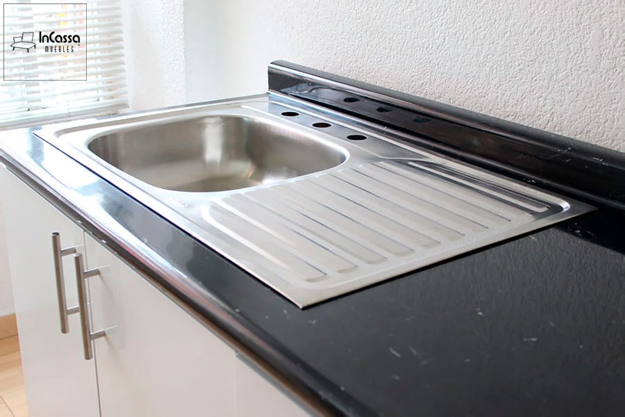 Una toma en ángulo que destaca la elegante tarja de acero inoxidable de la cocina modelo AUSTRIA, con escurridor integrado y cubierta de formaica en acabado mármol negro brillante. El mueble inferior, en color blanco brillante, incluye jaladeras metálicas modernas que contrastan con el diseño contemporáneo de la cocina. Ideal para mantener la limpieza y funcionalidad en tu espacio.