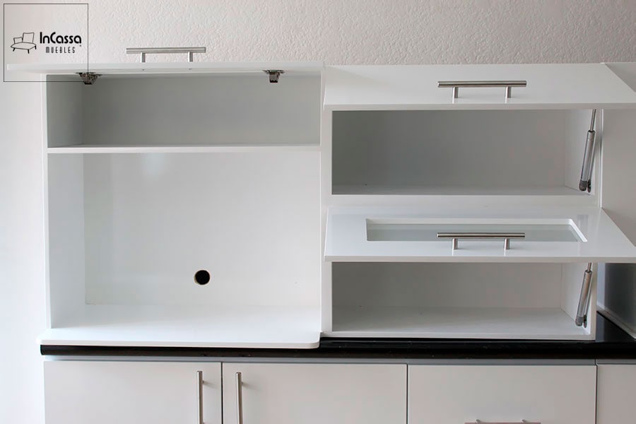 La imagen muestra una sección superior de la cocina AUSTRIA en color blanco, con puertas abatibles y pistones neumáticos. A la izquierda, el módulo con perforación trasera está diseñado especialmente para microondas. A la derecha, dos compartimentos con puertas abatibles de cristal esmerilado aportan un toque moderno.