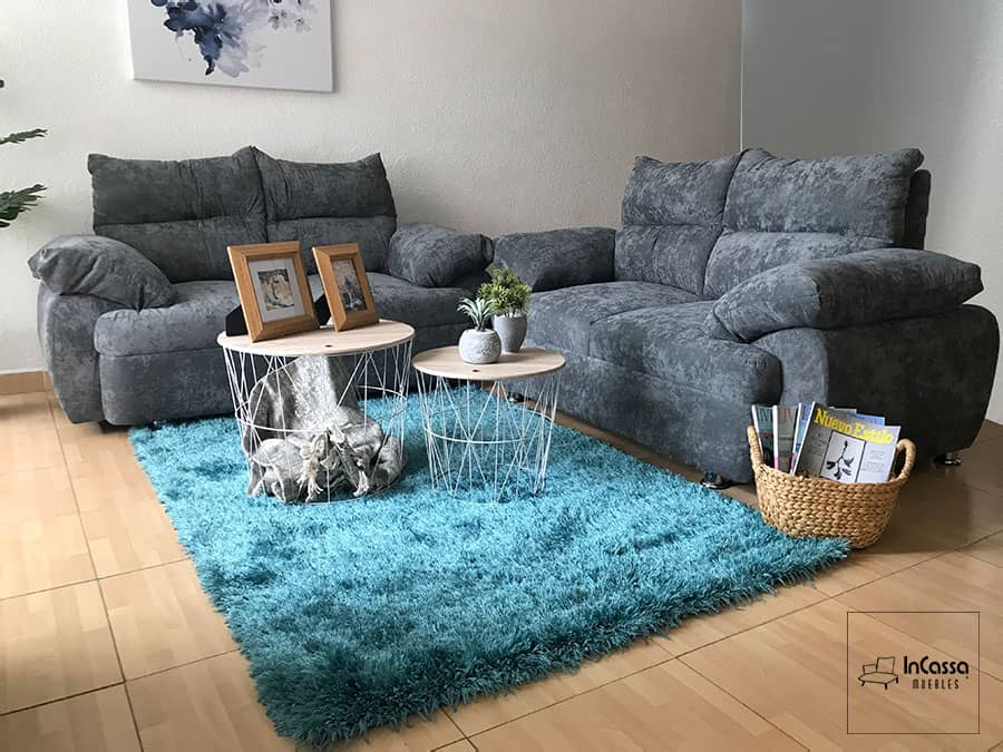 La foto presenta la Sala Modelo Bora Bora en su versión con dos love seat tapizados en suede gris oxford. El diseño moderno se realza con cojines amplios y brazos acolchados que ofrecen máximo confort, acompañados por una alfombra azul turquesa de textura suave y mesas circulares metálicas que complementan la decoración acogedora.