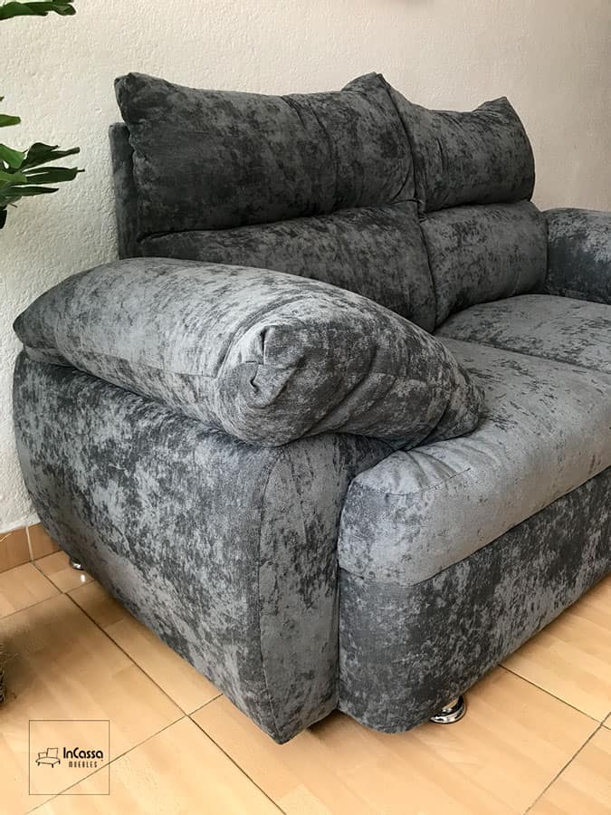 Una toma cercana muestra el acabado en suede gris oxford de la Sala Modelo Bora Bora. Se aprecian los brazos acolchados de gran volumen y el respaldo con cojines capitonados que garantizan comodidad. El tapizado suave y resistente aporta elegancia, mientras que las patas metálicas cromadas realzan su estilo moderno y sofisticado.