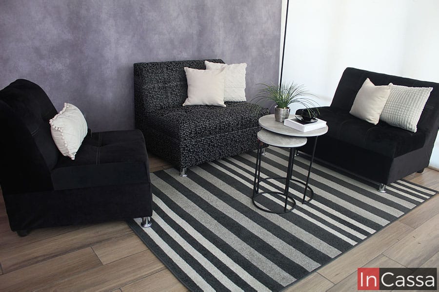 Sala minimalista instalada en una estancia con pared gris y piso de madera decorado con un gran tapete blanco con rayas negras. La sala está conformada por 3 módulos: un love seat y un sillón tapizados en suede negro, y un segundo love seat tapizado en lino estilo Da Vinci negro, cada módulo está adornado por cojines blancos.