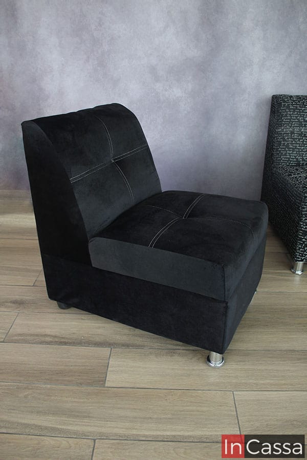 Se nos muestra con un mayor enfoque el sillón tapizado en suede negro, dándonos una mejor vista de su respaldo y asiento en los que resalta la textura de la tela, así como las costuras y el acabado capitoneado en el tapizado. Además, también podemos ver las patas que sostienen el módulo, siendo las delanteras metálicas mientras que las traseras son de plástico.