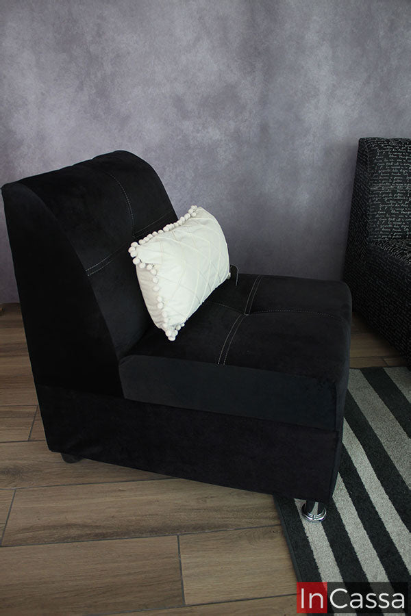 La imagen se concentra en mostrarnos el sillón de la sala, el cual esta tapizado en tela suede negro completo, en la que además se puede apreciar detalles como sus costuras y las patas que lo sostienen de las cuales las delanteras son de metal cromado mientras que las traseras son de plástico en color café. El mueble está decorado por un pequeño cojín blanco.