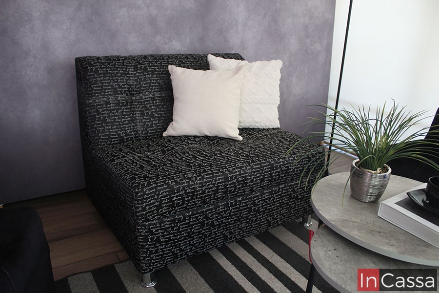 La foto destaca el love seat tapizado lino estilo Da Vinci negro, en la que también podemos ver sus patas metálicas delanteras; el mueble está instalado frente a una pared gris y presentado con un tapete blanco con rayas negras, además se encuentra adornado por dos cojines decorativos blancos.