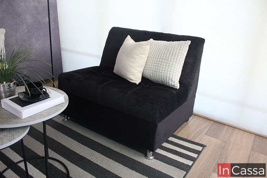 Una imagen que nos muestra uno de los love seat de la sala, tapizado completamente en suede color negro. El módulo esta instalado frente a una ventana de pantalla completa, junto a unas pequeñas mesas de centro grises con base metálica negra.