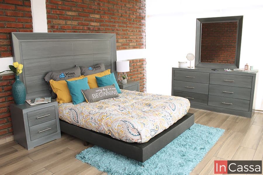 La imagen muestra la recámara Barcelona en elegante tono gris cenizo oscuro, con cabecera amplia, burós con jaladeras metálicas y tocador con espejo. Su diseño moderno resalta frente al muro de ladrillo, creando un ambiente acogedor y contemporáneo.