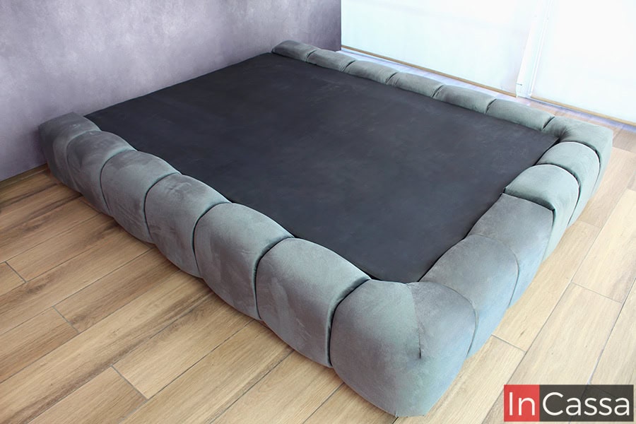 Una base de cama de estilo moderno y acogedor, tapizada en suave suede gris oxford, brindando una sensación de lujo y confort. Su diseño de bloques acolchonados a lo largo de los bordes la hace un elemento distintivo y elegante para cualquier dormitorio.