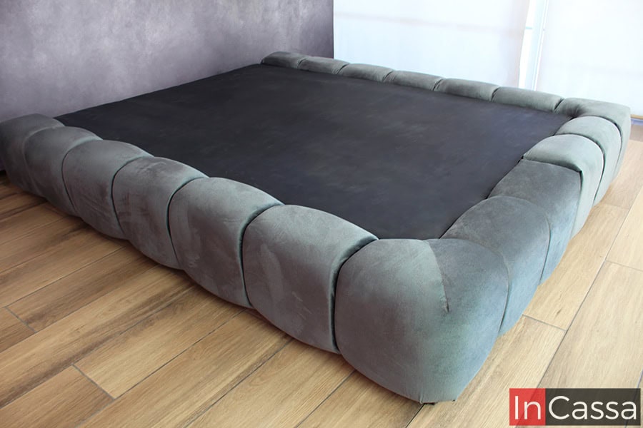 La base en suede gris oxford se presenta en una vista lateral completa, destacando su estructura baja y robusta, ideal para crear un ambiente relajado. El acolchado en todo el perímetro ofrece un toque de suavidad que complementa su diseño moderno.