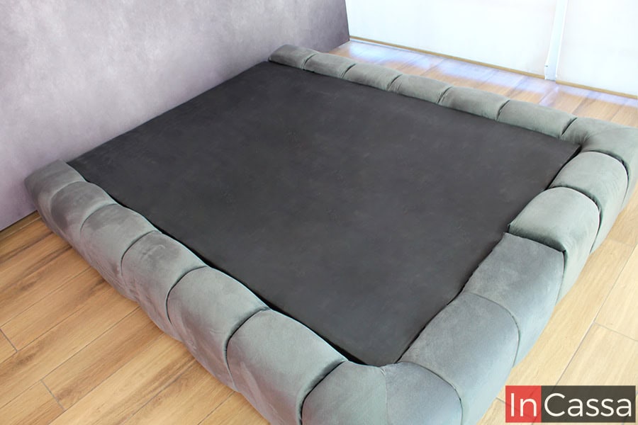 Con una vista desde arriba, se destaca la continuidad del diseño acolchado alrededor de la base tapizada en suede gris oxford, garantizando que el estilo envolvente se mantenga desde cualquier ángulo.