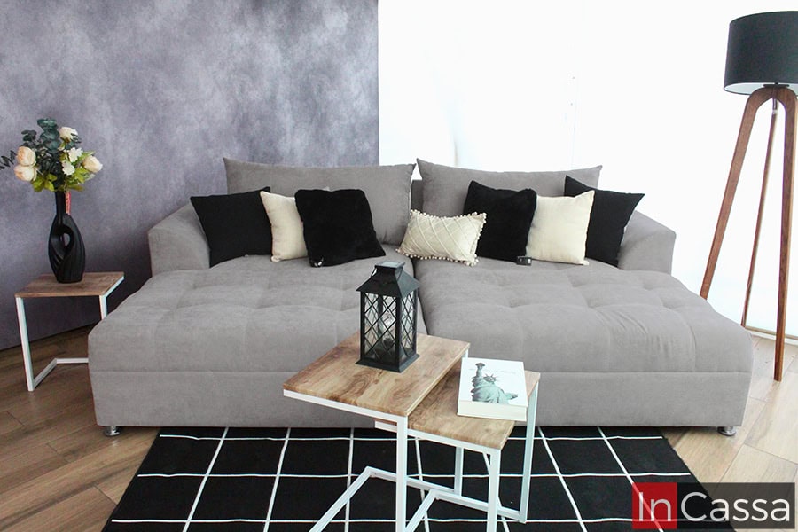 Una sala tipo cama de gran tamaño en tapiz suede gris perla, complementado con cojines en tonos contrastantes de negro, beige y crema. Su diseño elegante se combina con mesitas auxiliares de madera, un florero decorativo y una lámpara de pie moderna, creando un ambiente sofisticado.