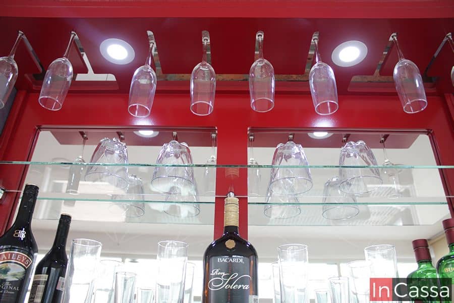 Primer plano de la cantina Bonn en rojo vivo, mostrando los soportes metálicos para copas suspendidas y la iluminación LED integrada en la parte superior. Los estantes de cristal aportan un estilo moderno y elegante que realza el ambiente.