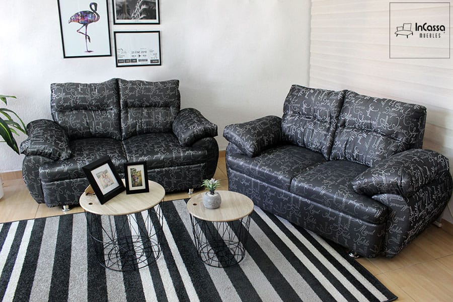 La imagen muestra la Sala Modelo Bora Bora en tapiz lino en estilo artistas negro con estampado de letras en gris, conformada dos love seat. Sus cojines amplios y brazos acolchados aportan comodidad, mientras que la alfombra a rayas y mesas circulares metálicas realzan su estilo moderno y acogedor.