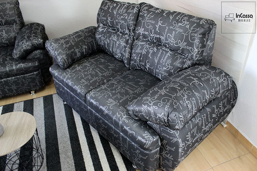 La foto presenta el love seat de la Sala Modelo Bora Bora, tapizado en lino artistas color negro con estampado de letras grises. Su diseño incluye respaldo alto con cojines capitonados y brazos acolchados que aportan máximo confort, destacando sobre la alfombra a rayas y la mesa circular decorativa.