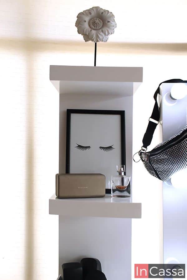 Se presenta un estante blanco con repisas decoradas con un cuadro de pestañas y perfume, añadiendo un toque artístico al espacio. Ideal para personalizar el tocador.