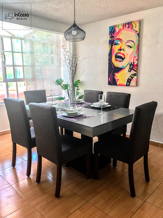 Una fotografía de ángulo amplio muestra un elegante comedor de seis sillas en acabado gris cenizo oscuro, acompañado de sillas tapizadas en tela lino color mouse con patas de madera de pino oscura. La luz natural realza el ambiente moderno del espacio.