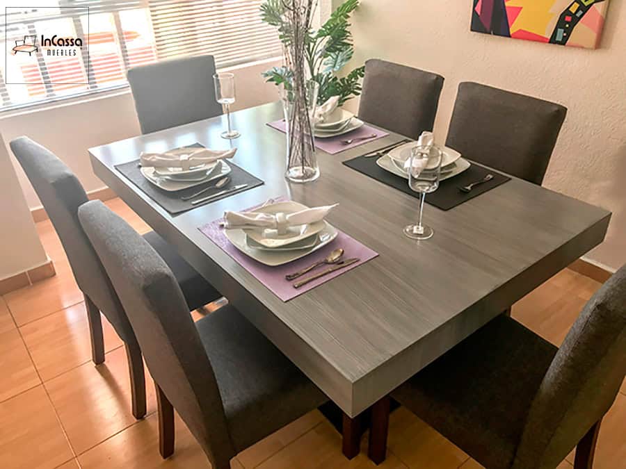 La imagen muestra una vista lateral superior de un comedor moderno para 6 personas, destacando la textura gris cenizo oscuro de la mesa y la elegante disposición de los utensilios de mesa. Las sillas tapizadas en tela lino color mouse complementan el conjunto con un estilo sobrio y contemporáneo.