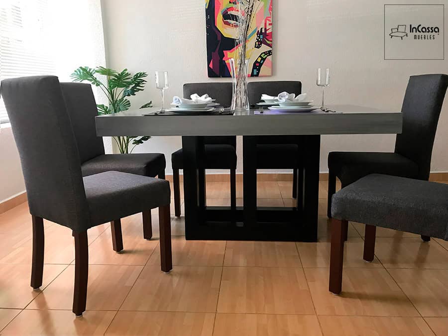 La fotografía presenta un ángulo frontal bajo del comedor de seis sillas, donde resalta la sólida base de pedestal color chocolate que sostiene la mesa de acabado gris cenizo oscuro. Las sillas tapizadas en tono mouse enmarcan el conjunto con un estilo moderno y elegante.