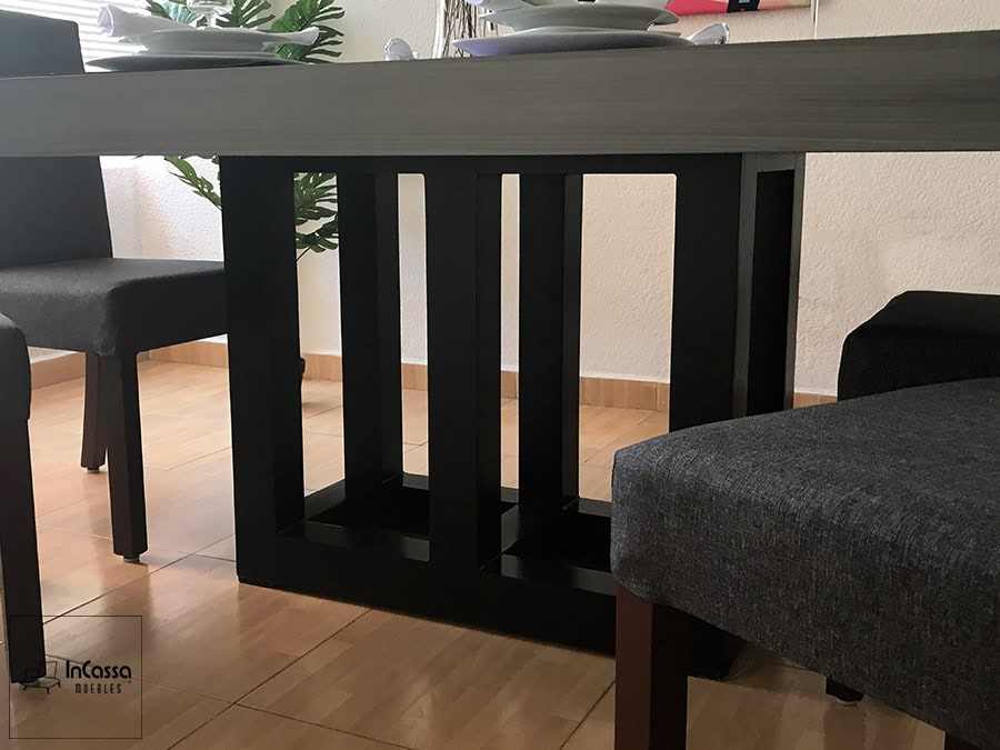 La imagen muestra un acercamiento a la estructura de pedestal del comedor, elaborada con un elegante diseño de figuras geométricas en acabado chocolate mate. Se aprecia la robustez de la base y parte de las sillas tapizadas en lino color mouse que complementan el conjunto.