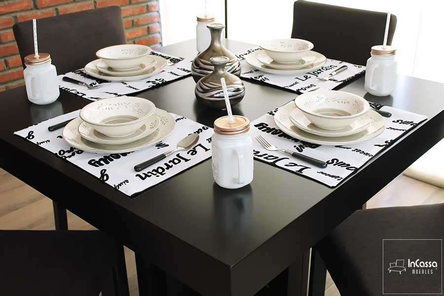 La imagen muestra una elegante mesa cuadrada de acabado chocolate, decorada con vajilla en tonos neutros y caminos de mesa blancos con tipografía negra. El ángulo superior resalta el diseño limpio y contemporáneo del comedor para cuatro personas.