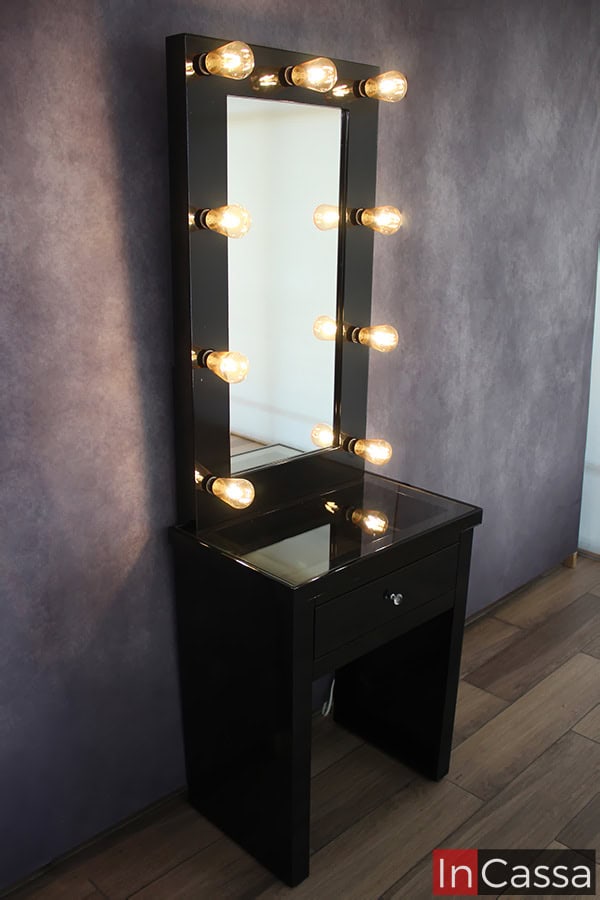Vemos el tocador desde una toma lateral, la cuál muestra de manera más completa la estructura total del mueble. Este se presenta en un acabado negro brillante en su totalidad, incluyendo un espejo con luces LED integradas cuya luz destaca la cubierta de cristal del tocador.