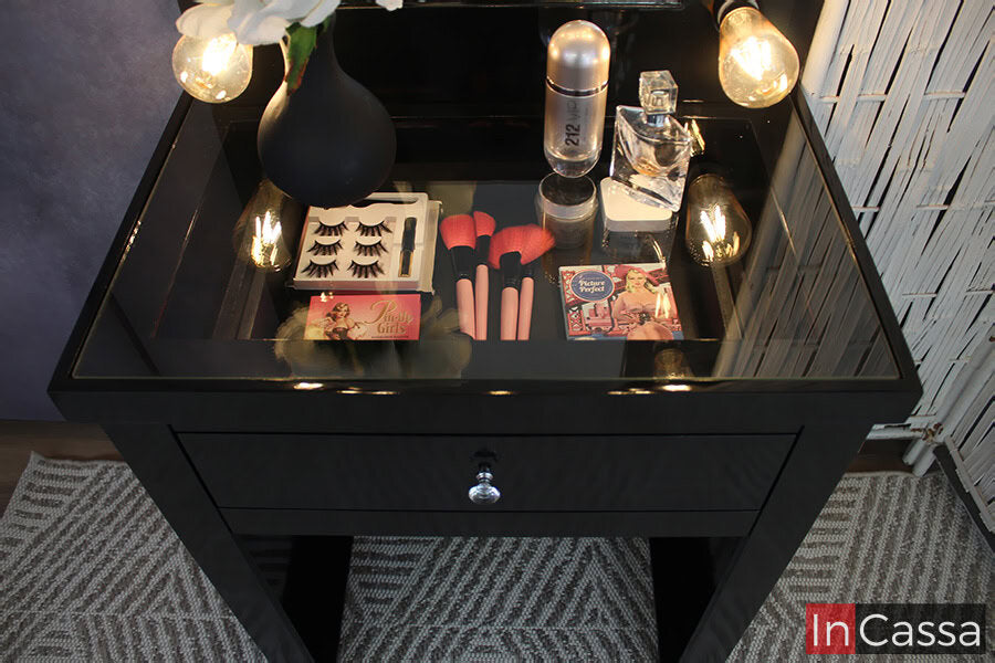 Una toma superior del tocador la cuál destaca su cubierta de cristal, con la cuál quedan a la vista los utensilios y maquillajes guardados en el cajón. También se puede apreciar la jaladera con forma de diamante incorporada al cajón, al igual que el acabado brillante de la pintura negra presente en todo el tocador.