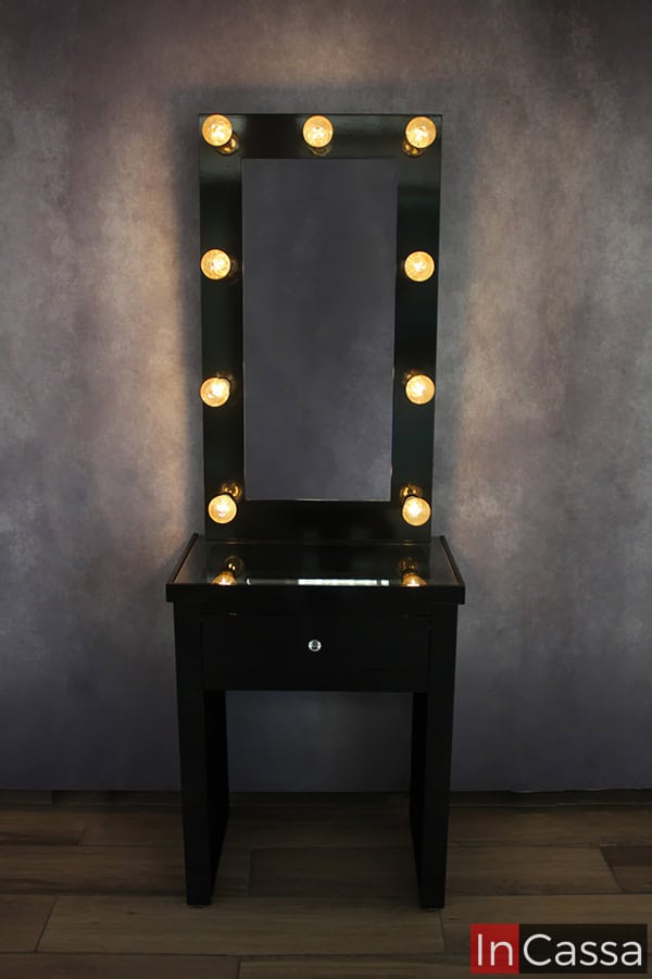 Foto del tocador desde una toma frontal, en la que podemos ver el mueble completo con sus luces LED encendidas, además de su cubierta de cristal. El tocador es exhibido en una presentación color negro brillante completo, instalado en una habitación con pared gris y piso de madera.