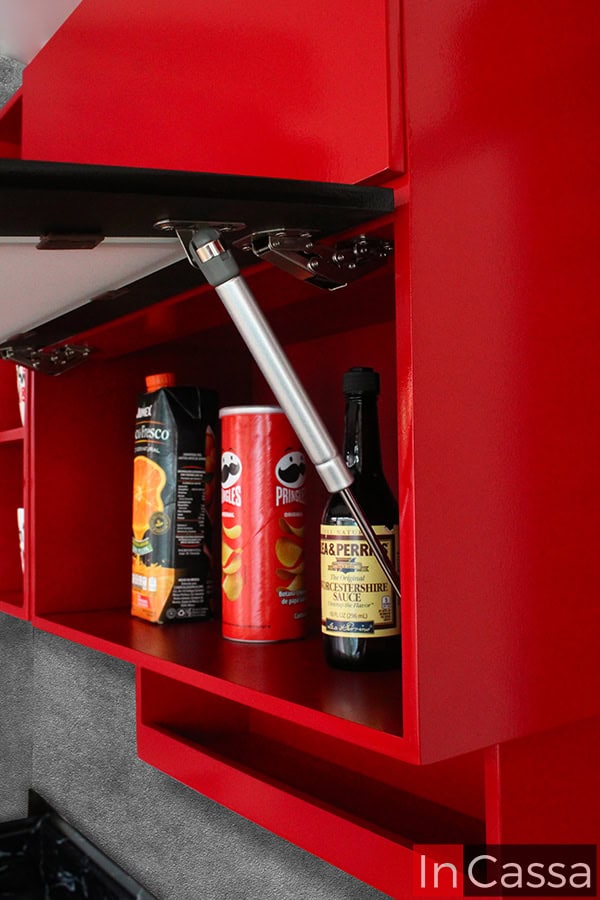 La imagen destaca el mecanismo de pistón neumático en los módulos superiores de la cocina integral. Este sistema asegura una apertura suave y silenciosa, facilitando el acceso a los compartimentos. El acabado interno combina perfectamente con el diseño vibrante en rojo, ideal para almacenar productos de manera organizada y a la vista.