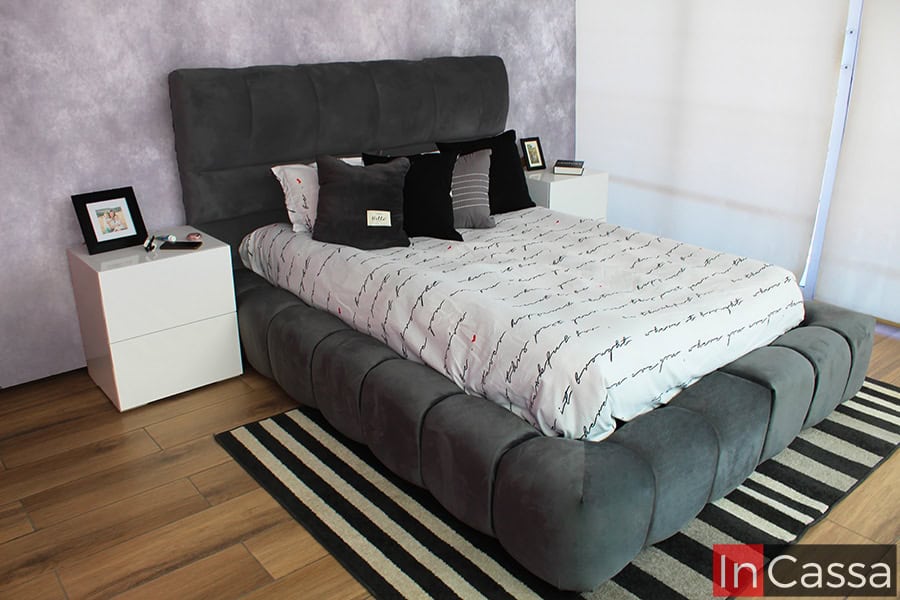 La foto muestra una cama tapizada en suave suede color gris oxford, acompañada de dos modernos buroes blancos con cajones de diseño minimalista. La cabecera acolchonada ofrece una sensación de confort envolvente, perfecta para cualquier dormitorio.