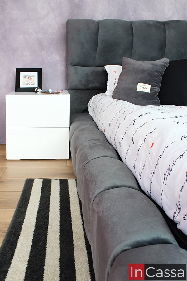 La imagen lateral captura el perfil de la cabecera y la base, destacando la estructura robusta pero elegante del conjunto. El contraste entre el tapiz suede gris oxford de la cama y los buroes blancos crea un equilibrio perfecto en la decoración.