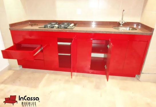 La imagen muestra la parte inferior de la cocina en acabado rojo vivo brillante, con puertas y un cajón completamente abiertos para revelar los amplios espacios interiores. La cubierta de formaica en estilo mármol negro integra una parrilla empotrada y tarja de acero inoxidable.