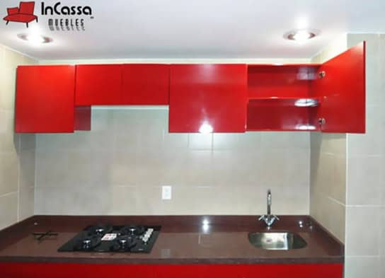Se presenta una vista frontal de las alacenas superiores en acabado rojo vivo brillante, con dos puertas abiertas que exhiben entrepaños interiores ideales para organizar utensilios o despensa. La cocina cuenta con cubierta de formaica en estilo mármol negro, parrilla empotrada y tarja.
