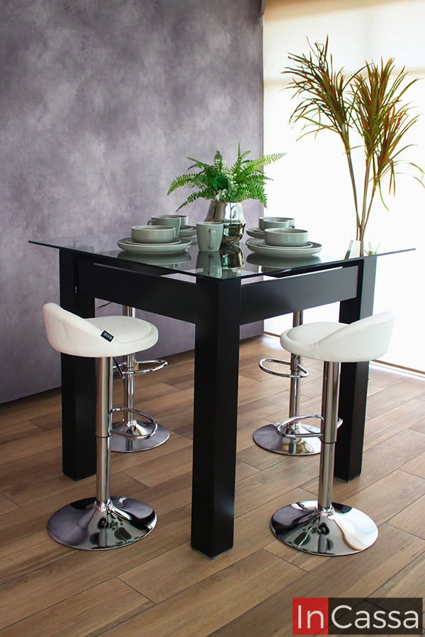 Moderno comedor alto en color chocolate con cubierta de cristal, acompañado de 4 bancos neumáticos tapizados en tacto piel blanco. El comedor se encuentra instalado en una estancia con pared gris y piso de madera.