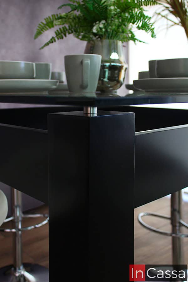 La toma se enfoca en la mesa del comedor alto, proporcionando una vista cercana de su base fabricada de MDF Chileno en acabado chocolate mate. Además también se pueden apreciar los separadores metálicos que permiten una posición suspendida en la cubierta de cristal de la mesa.