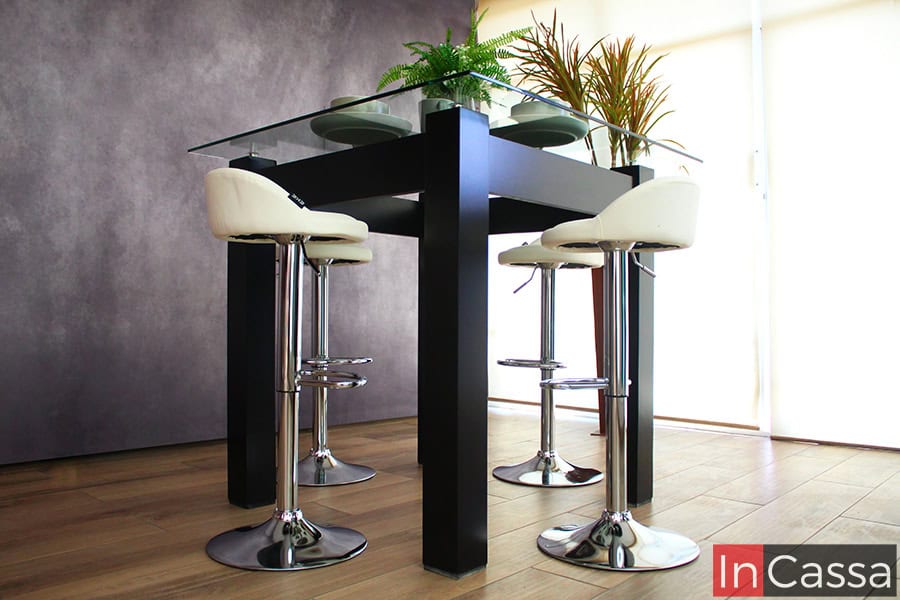 La fotografía se enfoca en la base de MDF del comedor resaltando el alto de sus patas al igual que su acabado chocolate mate. La toma también permite apreciar mejor la cubierta de cristal del comedor así como sus bancos neumáticos con asientos de tacto piel blanco.