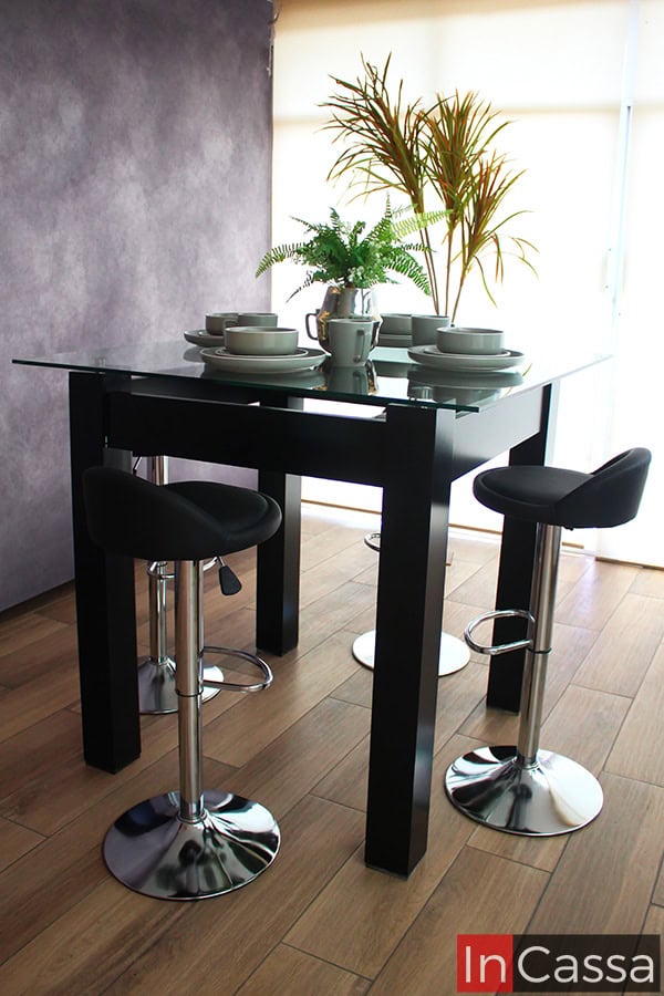 La foto muestra una toma completa del comedor alto en color chocolate mate con 4 bancos neumáticos tapizados en tacto piel negro; además, la cubierta de cristal del comedor queda resaltada gracias a la iluminación natural proporcionada por la ventana completa de la estancia.