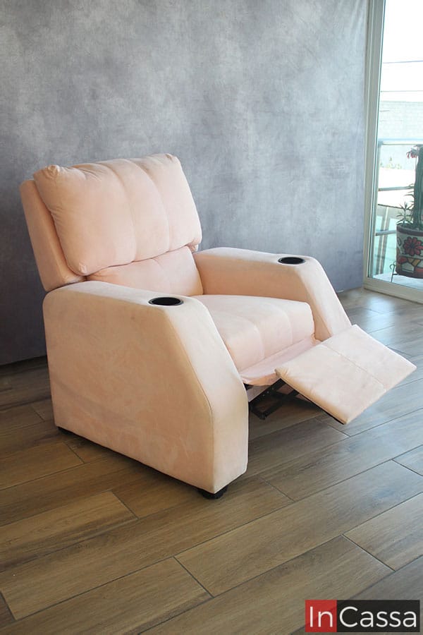 En esta toma lateral vemos el sillón de tapiz suede rosado en una posición levemente inclinada, en la que se puede observar parte de su mecanismo gracias a que el reposapies se encuentra extendido. Adicionalmente, también son visibles los porta vasos incorporados a los reposabrazos del sillón.