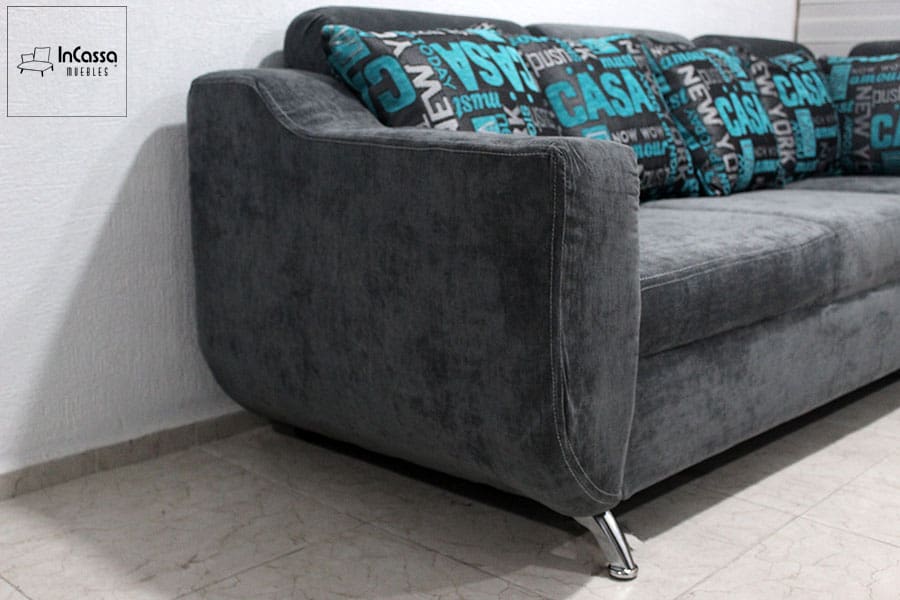 Reposabrazos lateral de la sala esquinera de suede gris con patas metálicas cromadas y cojines decorativos turquesas. Su diseño moderno y resistente es ideal para complementar cualquier sala de estar con un toque de estilo contemporáneo.
