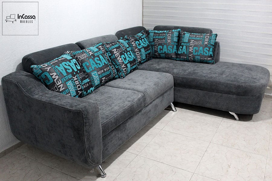 Sala esquinera tapizada en suede gris con chaise long y cojines decorativos de letras turquesa. Ideal para espacios contemporáneos, combinando funcionalidad y diseño moderno para un ambiente acogedor y elegante.