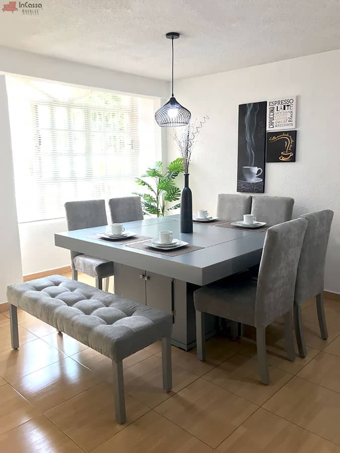 Una fotografía amplia muestra el comedor Cambridge para 8 personas en color silver completo, compuesto por seis sillas y una banca tapizada en suede gris perla. La mesa central con base tipo gabinete destaca en un entorno luminoso y armonioso con decoración moderna