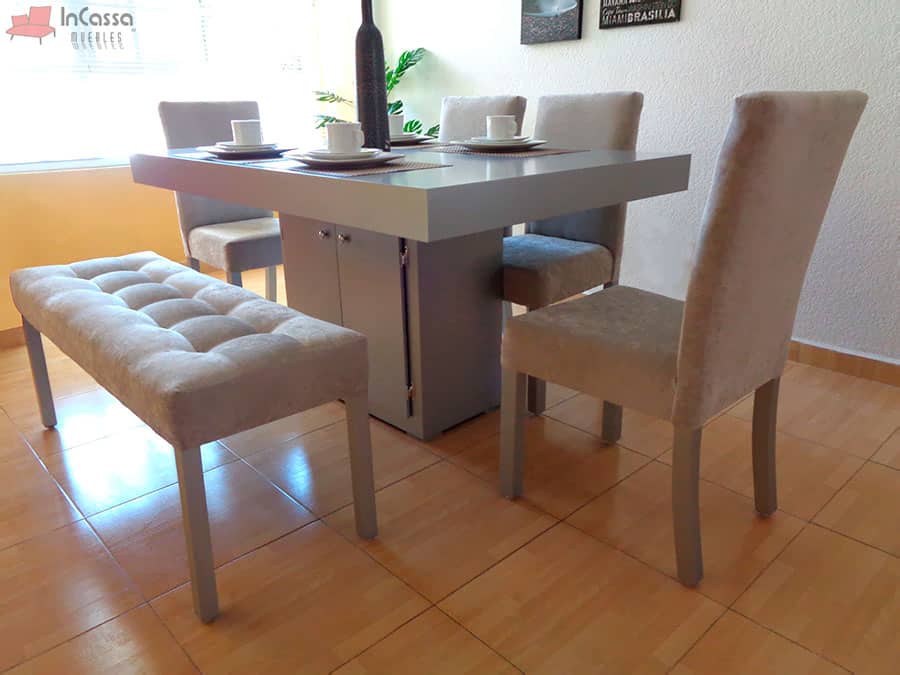 La foto presenta el comedor Cambridge para 6 personas desde un ángulo lateral, resaltando su mesa de cubierta gruesa y color silver completo, gabinete con puertas centrales y sillas tapizadas en suede gris perla. La banca capitonada completa el conjunto con un estilo acogedor.