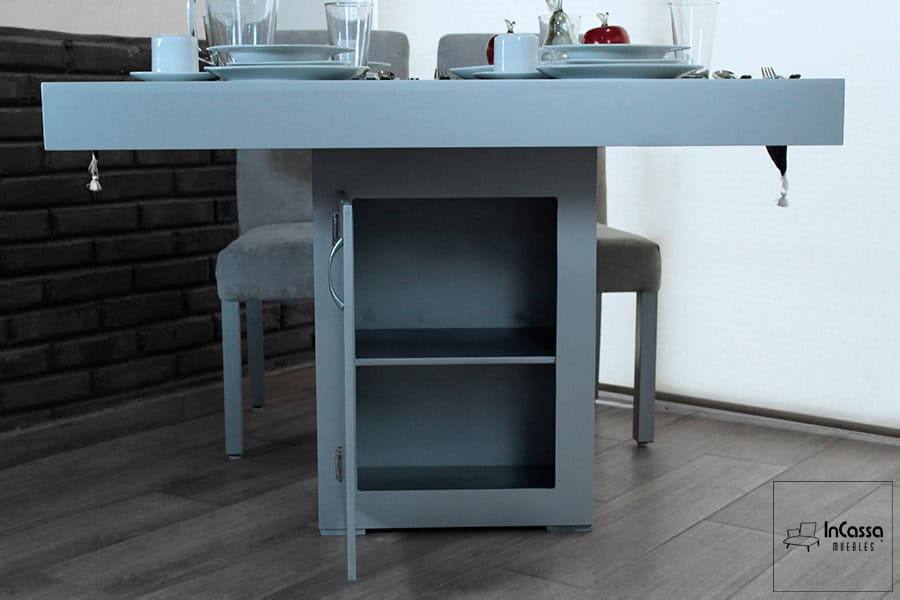 La imagen muestra el gabinete integrado en la base de la mesa del comedor modelo Cambridge para 4 personas, con puerta abierta que revela una repisa interna, todo en acabado silver completo. Ideal para almacenar manteles, utensilios o elementos decorativos de uso diario.