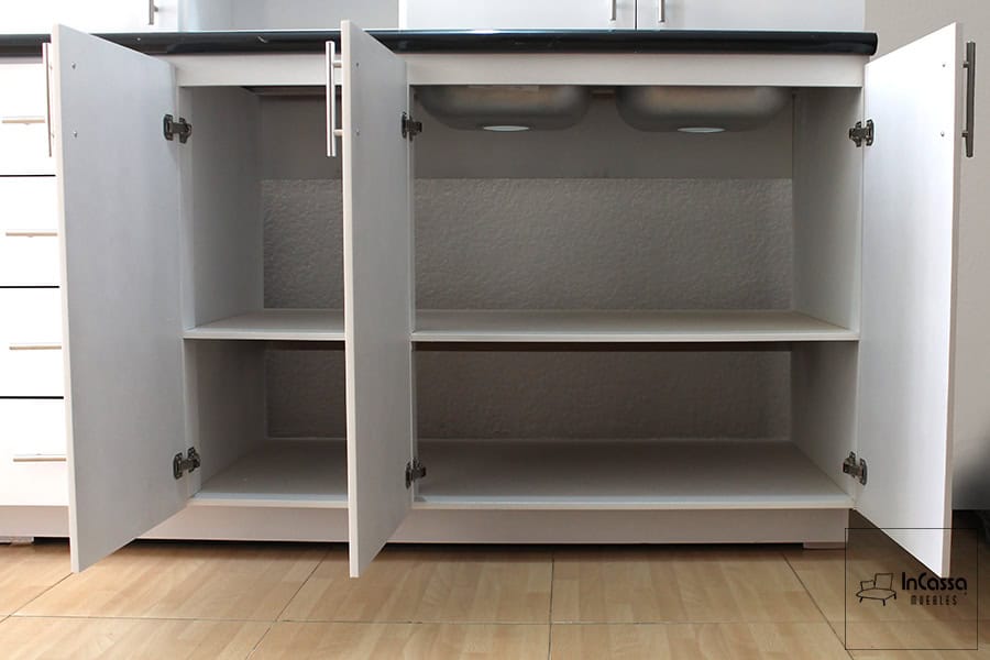 La foto muestra el módulo inferior del mueble de cocina en MDF blanco, con cuatro puertas abatibles abiertas que permiten observar su interior dividido por una repisa. Se aprecia la parte inferior de una tarja doble de acero inoxidable y la cubierta superior en acabado mármol negro. Las jaladeras metálicas rectas y las bisagras de excelente calidad completan los detalles visibles desde este ángulo.