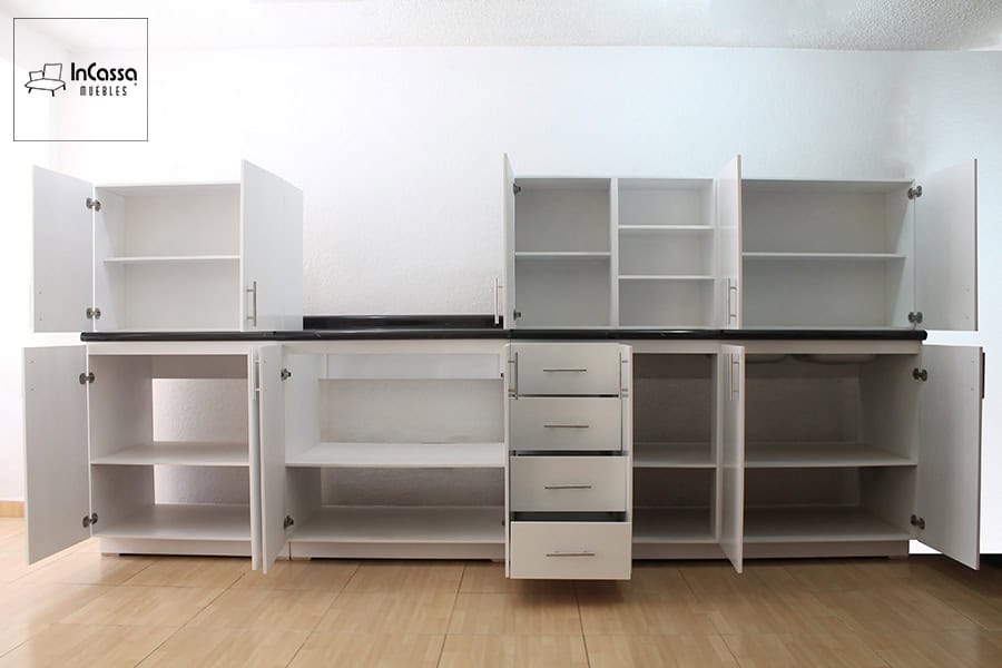 La imagen presenta una vista directa del modelo de cocina integral en color blanco, con todas sus puertas y cajones abiertos para revelar la organización interna. Se aprecian múltiples estantes funcionales, cuatro cajones centrales con correderas metálicas y módulos superiores con repisas abiertas. La cubierta de formaica en acabado mármol negro realza el contraste moderno del diseño.