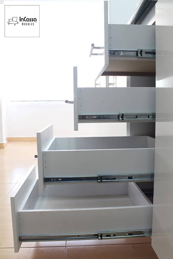 La imagen presenta los cuatro cajones del módulo inferior totalmente abiertos, mostrando su estructura en MDF blanco con correderas metálicas telescópicas de extensión completa. Las jaladeras rectas de acero inoxidable aportan un detalle moderno y funcional. Esta vista en perspectiva permite apreciar la capacidad de almacenamiento y la calidad de los herrajes empleados.
