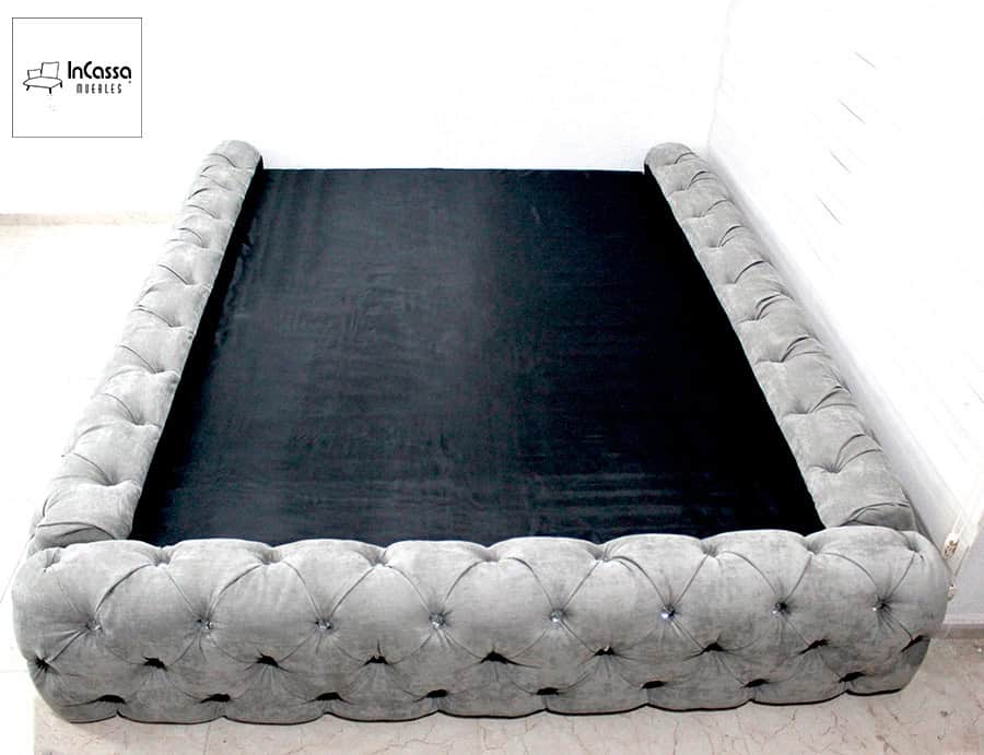 Base tapizada en tela suede gris perla con acabado capitoneado, instalada frente a una pared blanca.